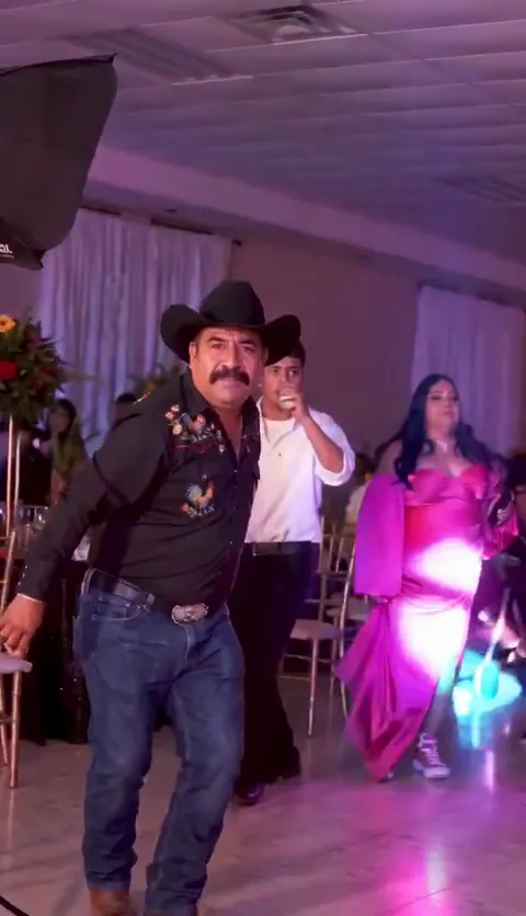 A man with a moustache in a black cowboy hat and embroidered shirt staring into the camera at a party