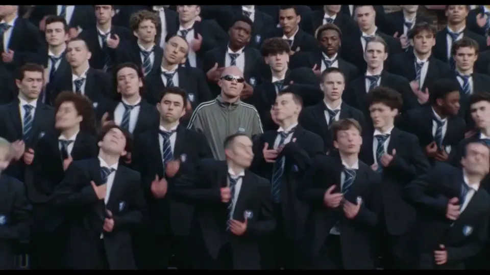 Center figure in a grey tracksuit standing amid a dark-suited dancing crowd